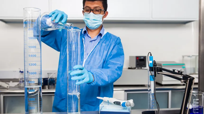 Laboratory analyst in a blue coat, mask and gloves pouring water from a flask into a graduated cylinder beside a pH meter
