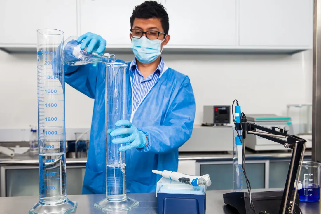 Laboratory analyst in a blue coat, mask and gloves pouring water from a flask into a graduated cylinder beside a pH meter and an electronic pipette