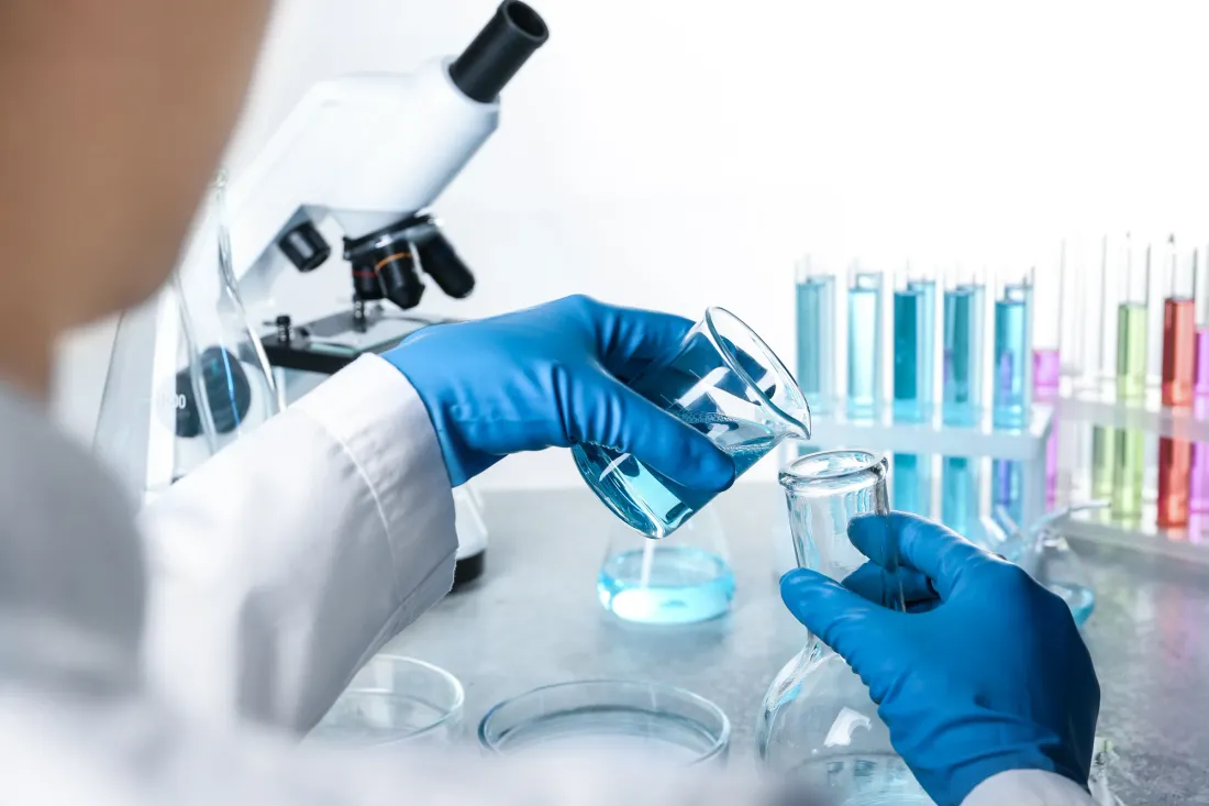Gloved hands pouring blue liquid from a beaker into a flask beside a microscope and racks of coloured test tubes