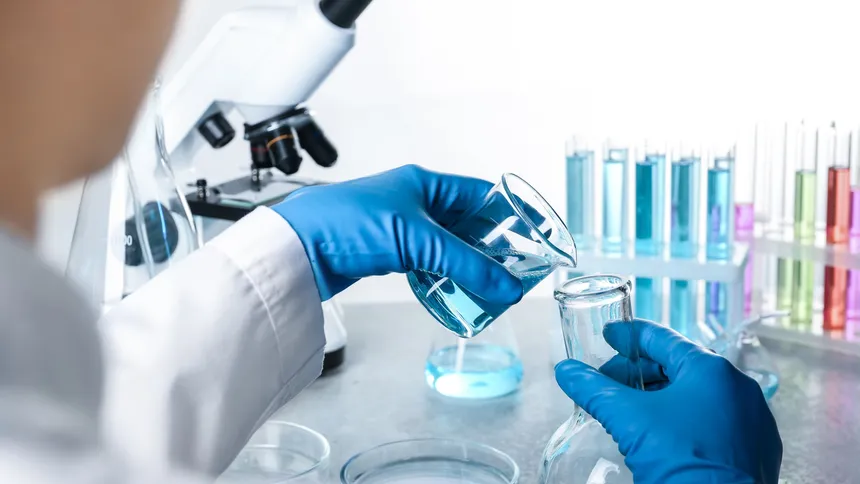 Gloved hands pouring blue liquid from a beaker into a flask beside a microscope and racks of coloured test tubes