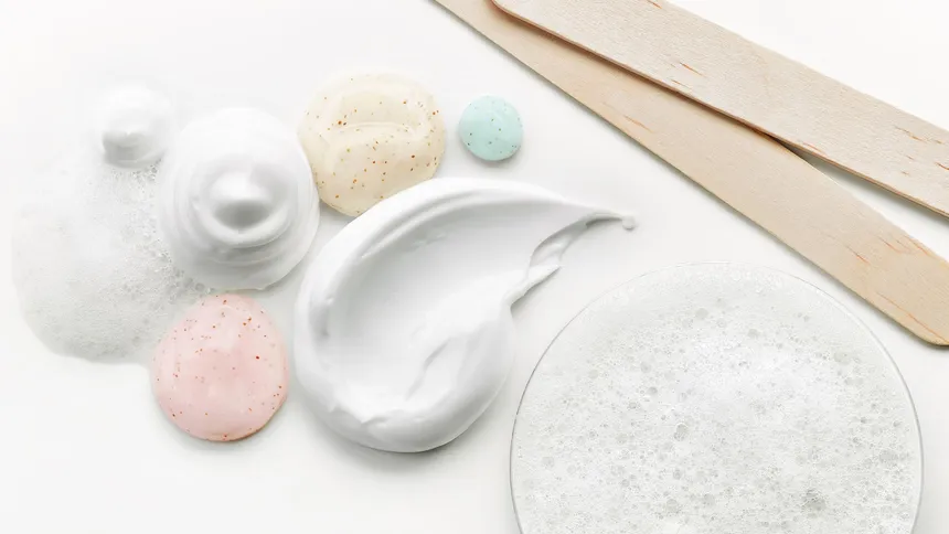 White creams, foam and speckled pink, beige and blue gels beside wooden spatulas on a white surface