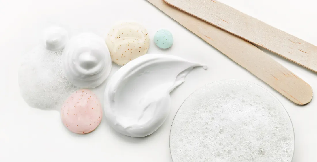 White creams, foam and speckled pink, beige and blue gels beside wooden spatulas on a white surface