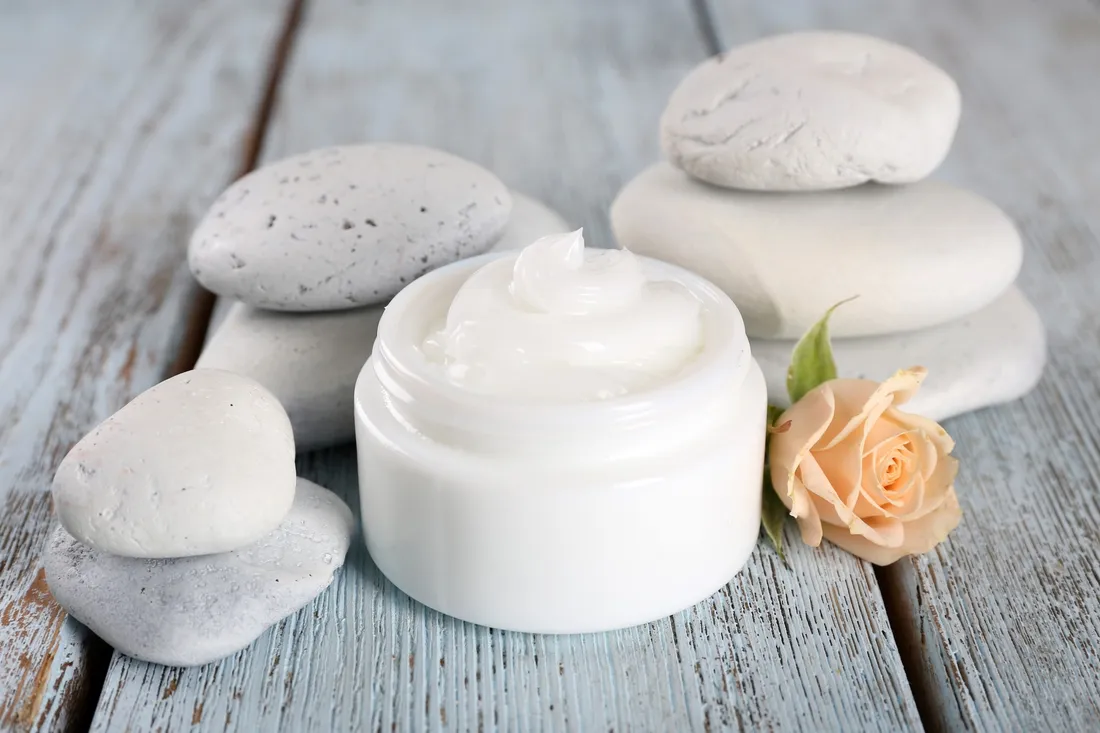Open jar of white cream beside smooth white stones and a peach rose on a pale blue wooden table