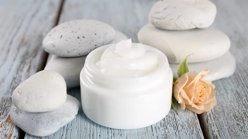 Open jar of white cream beside smooth white stones and a peach rose on a pale blue wooden table