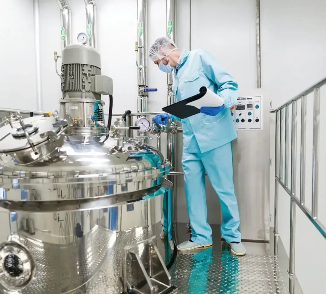 Operator in a gown, hairnet and mask checking a stainless steel process tank with a clipboard beside its control panel