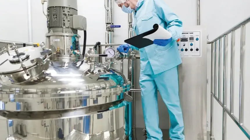 Operator in a gown, hairnet and mask checking a stainless steel process tank with a clipboard beside its control panel