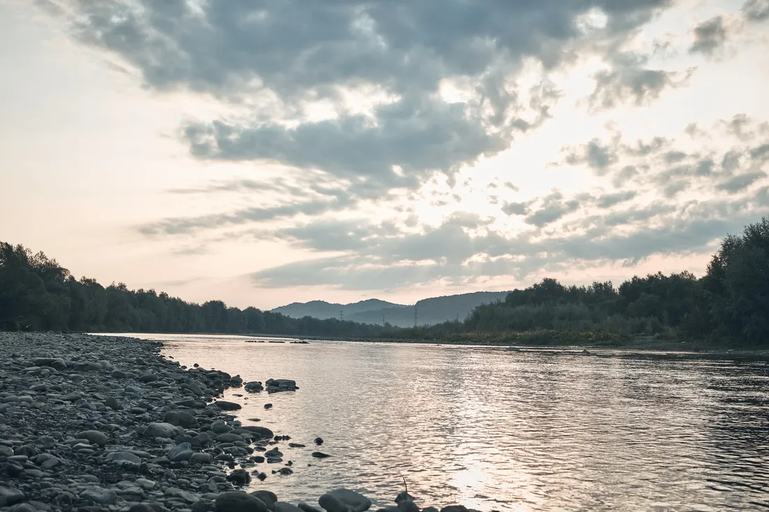 River with a stony bank flowing between wooded hills under a cloudy sky
