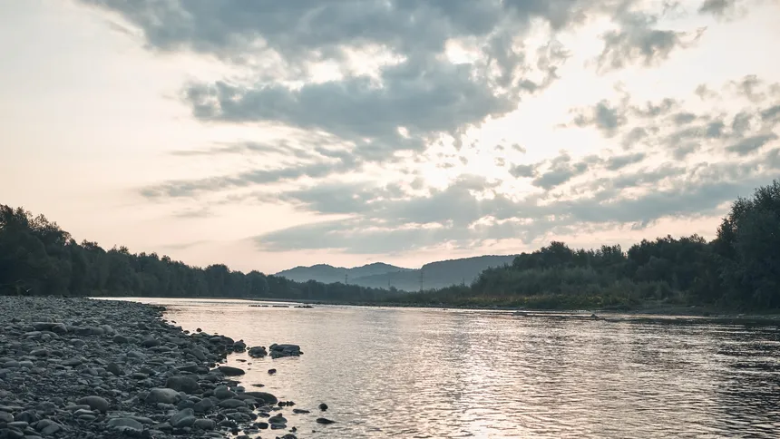 River with a stony bank flowing between wooded hills under a cloudy sky