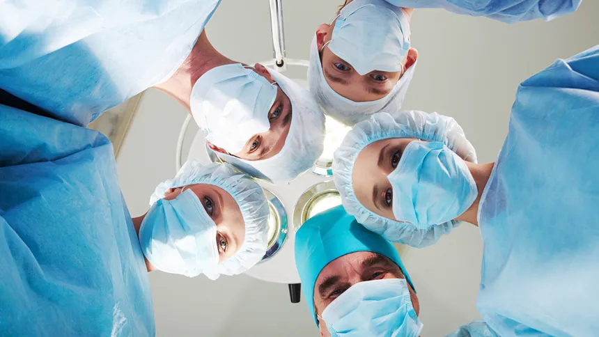 Five surgical staff in blue gowns, caps and masks looking down into the camera beneath an operating theatre lamp