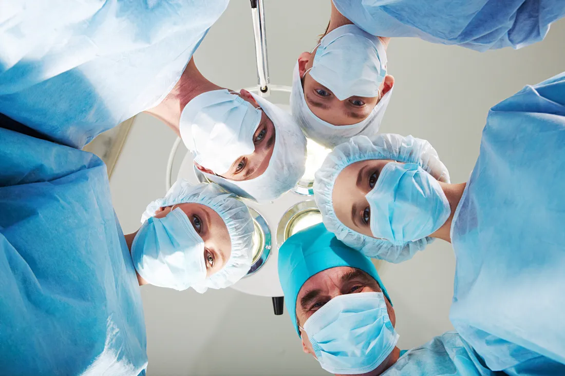 Five surgical staff in blue gowns, caps and masks looking down into the camera beneath an operating theatre lamp